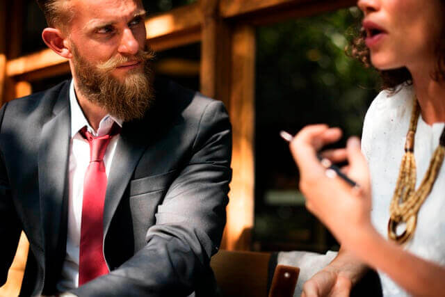 Man with a beard in a suit listening closely to someone else.